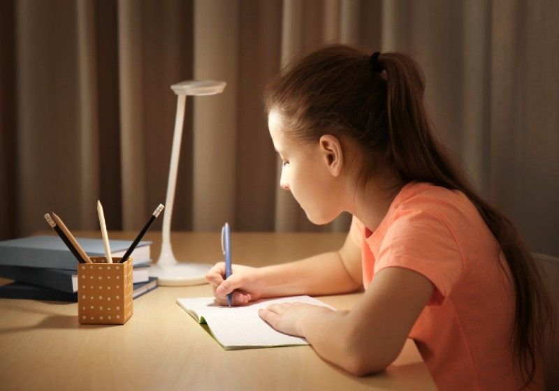 Menina estudando sobre a mesa com iluminação de um abajur